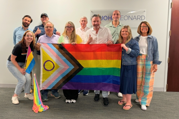 JOHNLEONARD team members standing together indoors holding a Progress Pride flag and a small Intersex-Inclusive Pride flag, celebrating Pride.