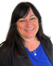 Headshot of a smiling woman with long dark brown hair, wearing a blue blouse and black sweater against a white background.